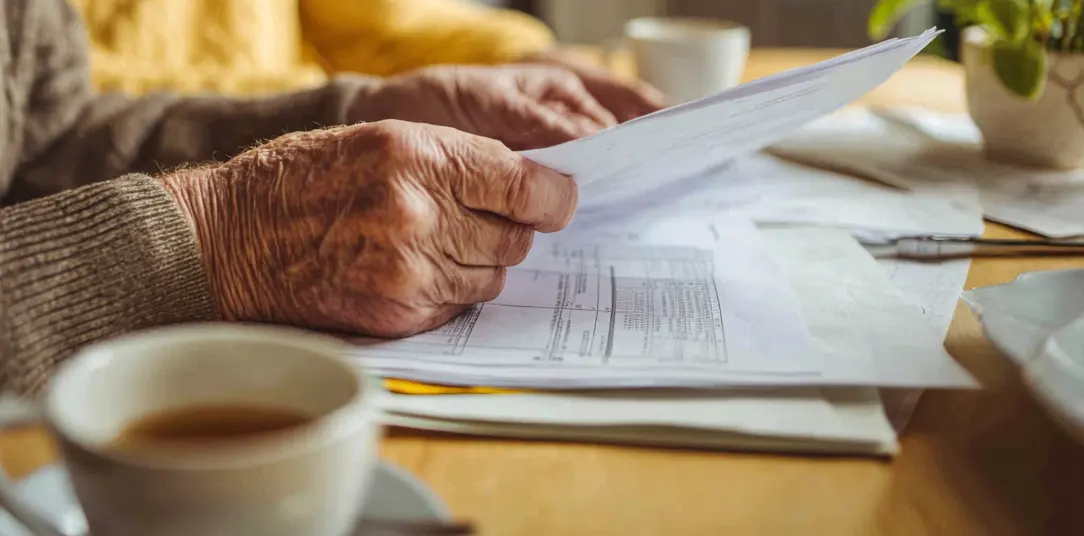 Elderly hands sorting through paperwork at a kitchen table beside a cup of tea